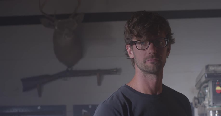 A man stands near a rifle and deer head mounted on the wall