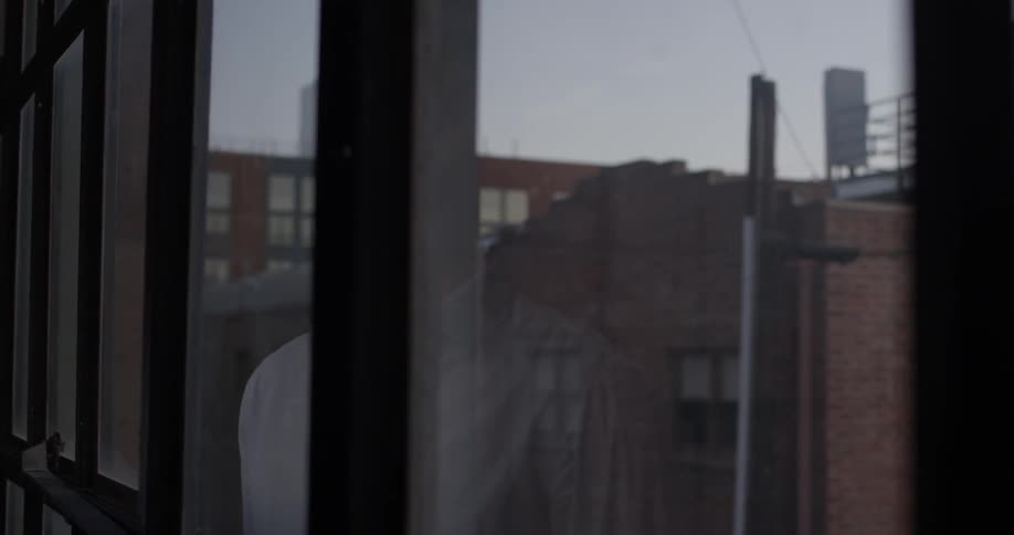 A man in a white shirt looks out a window at the city