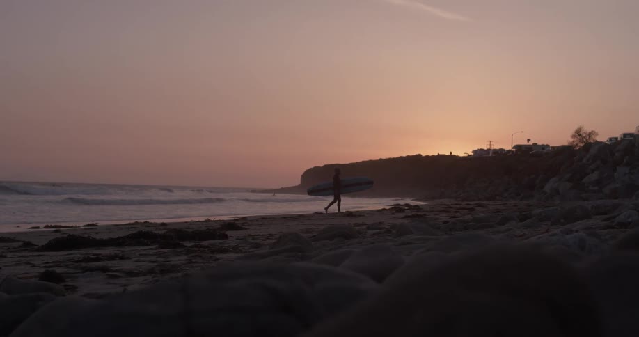 Surfer carries surfboard while walking on the beach at sunset