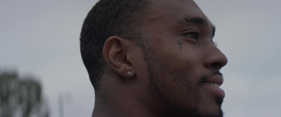 A man with a cross tattoo under his eye smiles as he looks into the distance