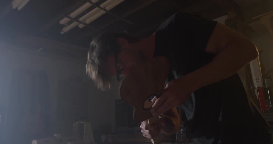 A man blows sand of a piece of a ukulele then continues sanding it