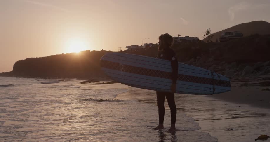 A man wearing a wet suit stands next to the ocean holding his surfboard at sunset