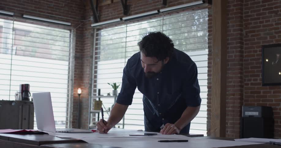 A man makes a note on a paper among many other papers while looking at a tablet