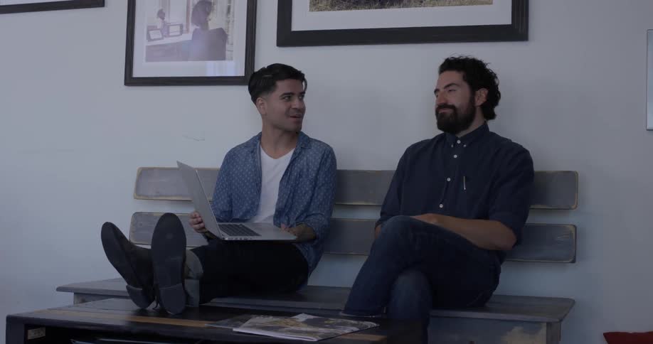 Two men are sitting on a bench chatting with each other while one holds a laptop