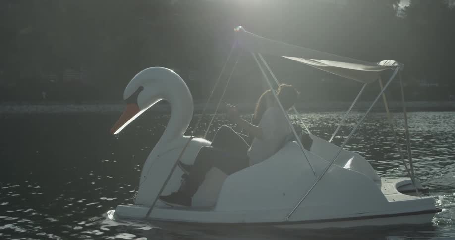 A man and women pedal a boat shaped like a swan through a lake in the sunshine