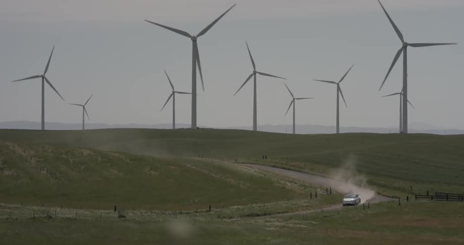 Windmills or wind turbines can be seen dotting the horizon in large numbers as a white sedan car rolls through the countryside road creating dust