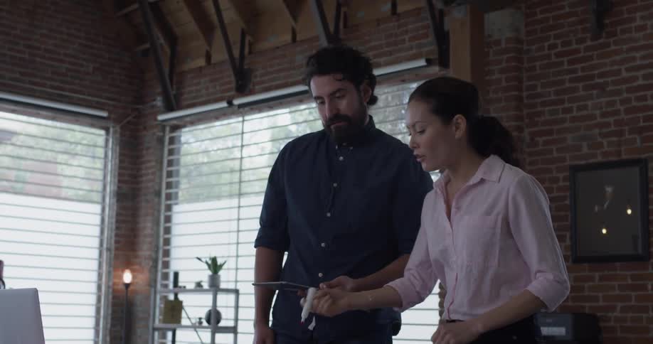 A man and a woman discuss things on a computer screen as well as a tablet screen and the women writes on some papers