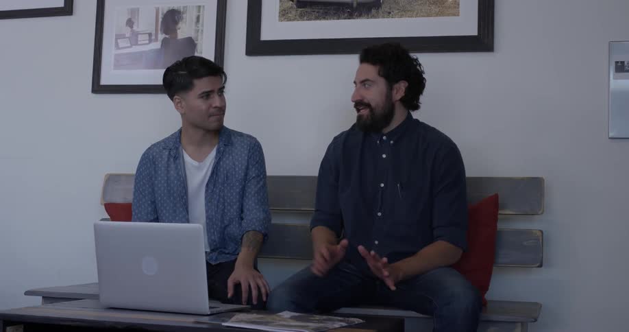 Two men have a discussion on a bench indoors in front of a laptop