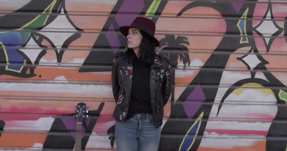 A woman leans against a garage door with graffiti on it and her guitar is to her right side