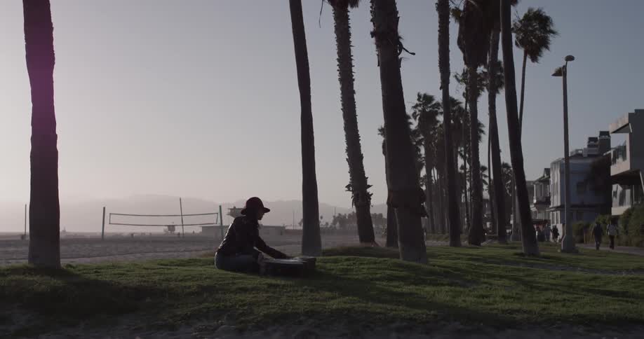 A woman walks to a place in the grass between some trees carrying a guitar then sits down and begins to play the guitar