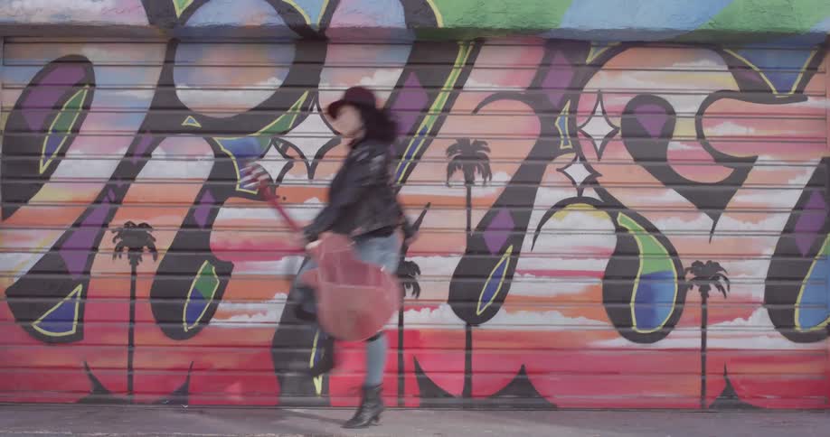 A woman with a guitar skips across a wall with graffiti on it