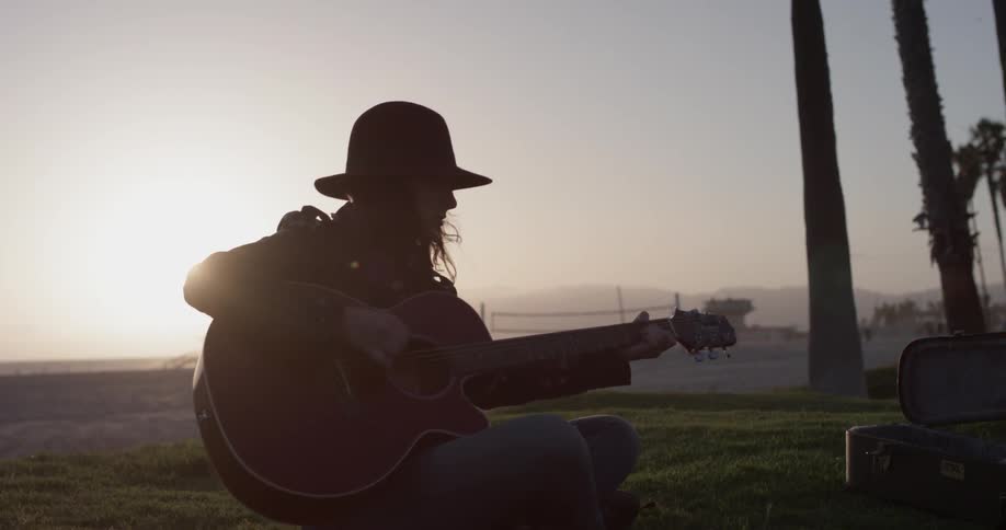 A woman sits in the sun playing guitar as someone rides by on a bike
