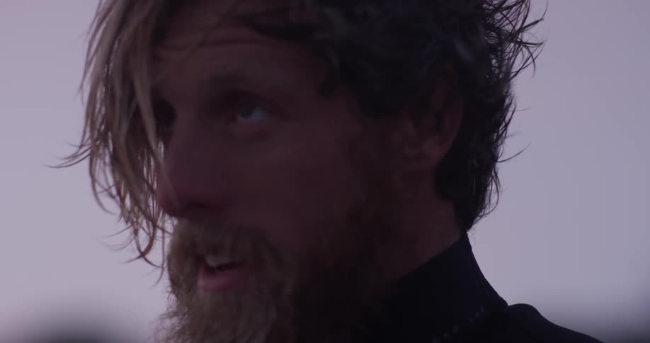 A man stands and smiles after the sun has set a bit with a beard and long blond hair wearing a wet suit outside at the beach