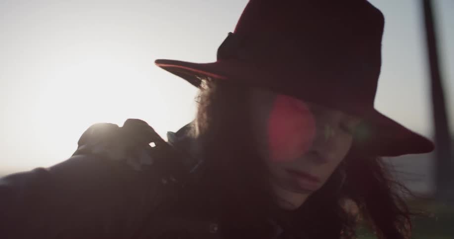 A woman with long hair plays guitar outside on a cloudy day while wearing a jacket and hat