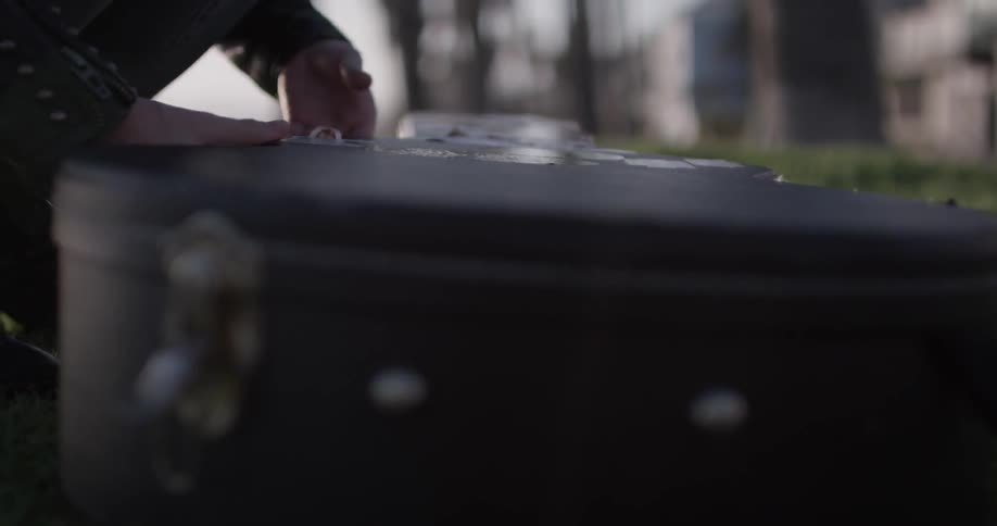 A woman sits down at the park and unpacks her guitar then holds it to begin playing