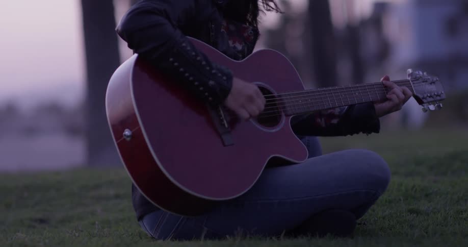 A woman with a flat brimmed hat strums her guitar as she sits on the grass at dusk