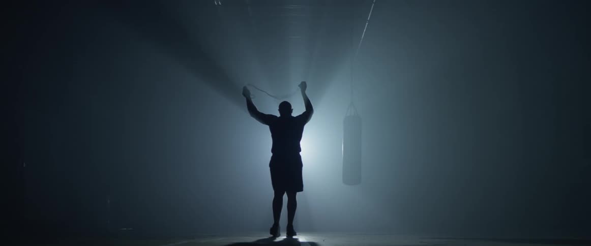 A backlit muscular man walks into a gym and uses a jump rope