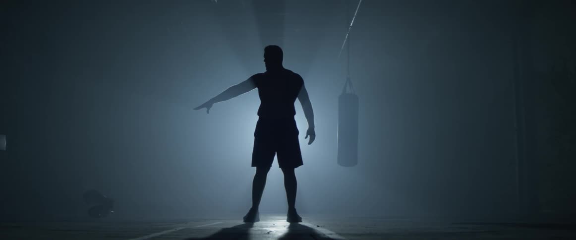 A muscular man stands in front of a light and throws boxing gloves to his right
