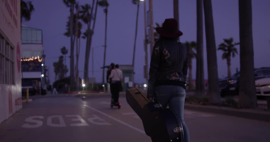 A woman carrying a guitar in a case walks down a city street after sunset