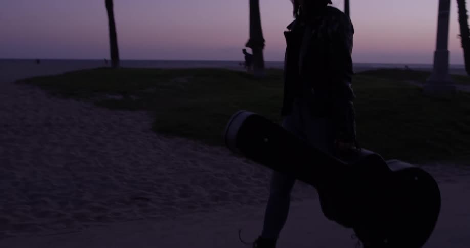 Woman with guitar case walks down street with palm trees near the beach at dusk