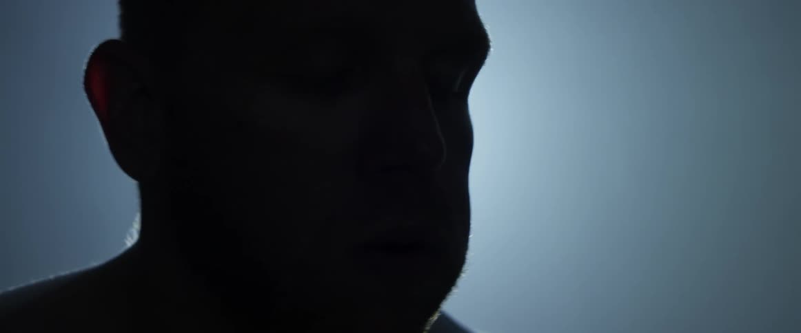 Shadowy man with no shirt stands in dim room