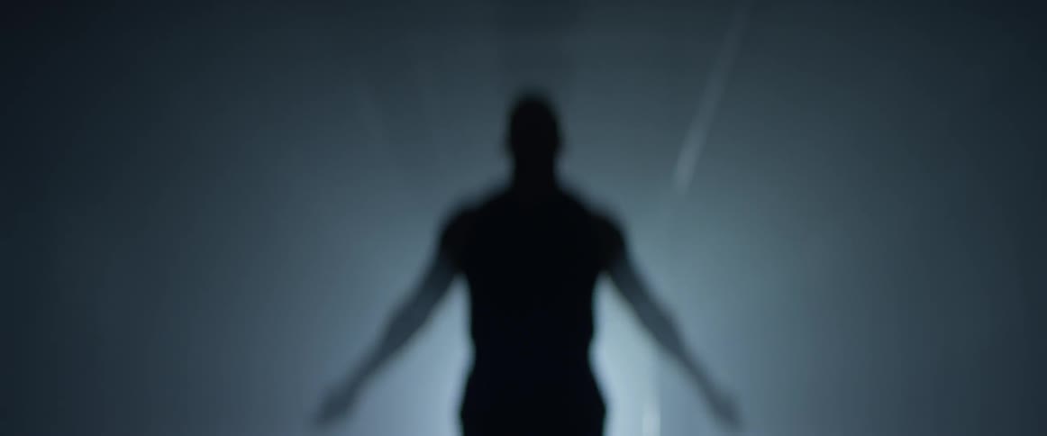 A man in a dim lit room eclipses the only light shining and jump ropes in front of it