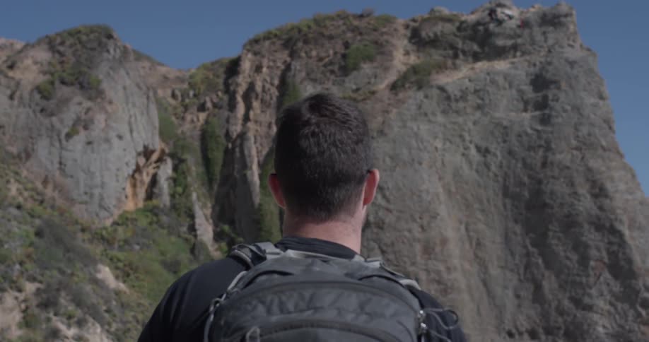 A man with a backpack on stares at a cliff under a clear blue sky