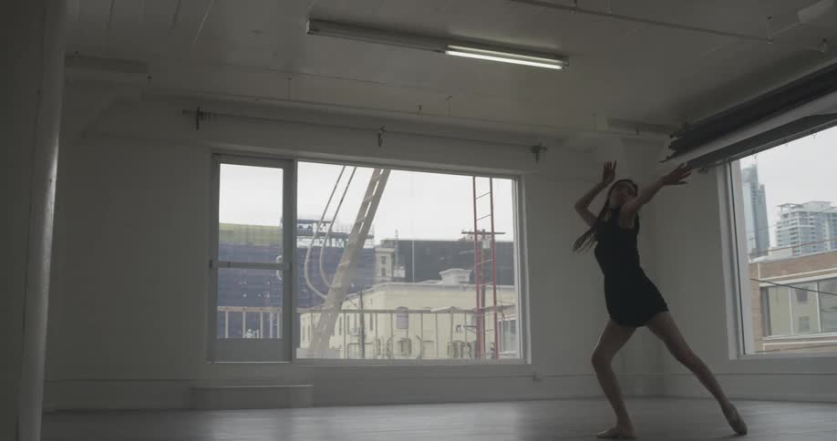 A woman with long hair and a black dress is dancing in slow motion inside a studio in the city