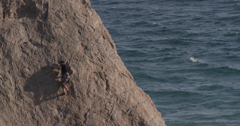 A man scales a tall rock above a body of water without much climbing equipment