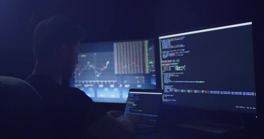 A man typing on a laptop with two large monitors behind it