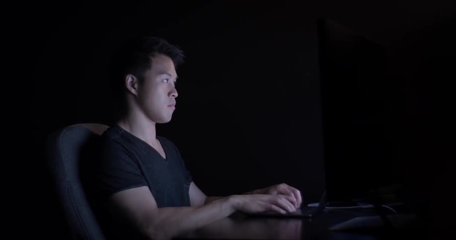 A man sitting at a desk typing on a laptop keyboard in a dark room