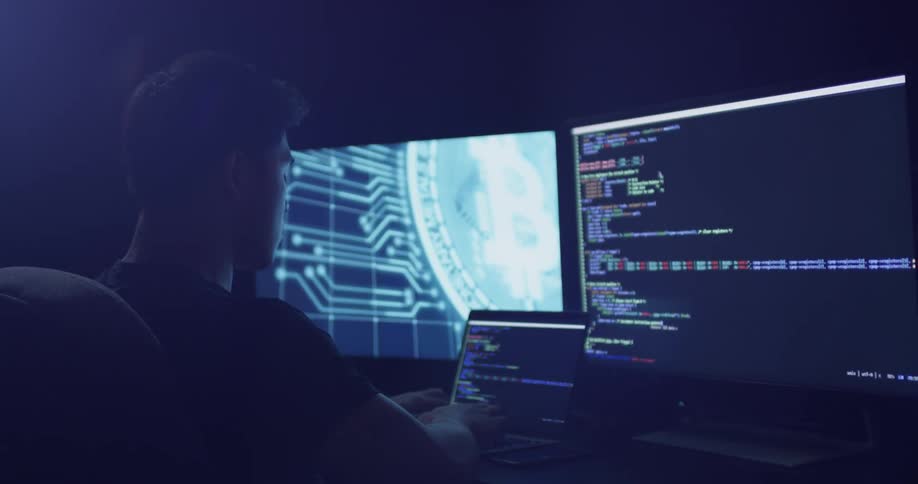 Man types on a laptop sitting in front of two monitors