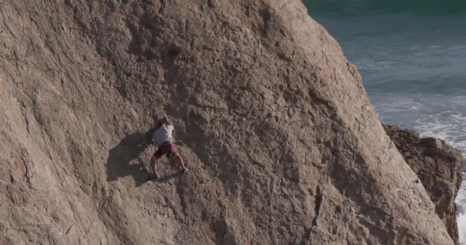 A man rock climbs onto a beachside rock at midday