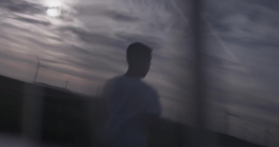 A man can be seen in the reflection of a car window standing under a cloudy sky