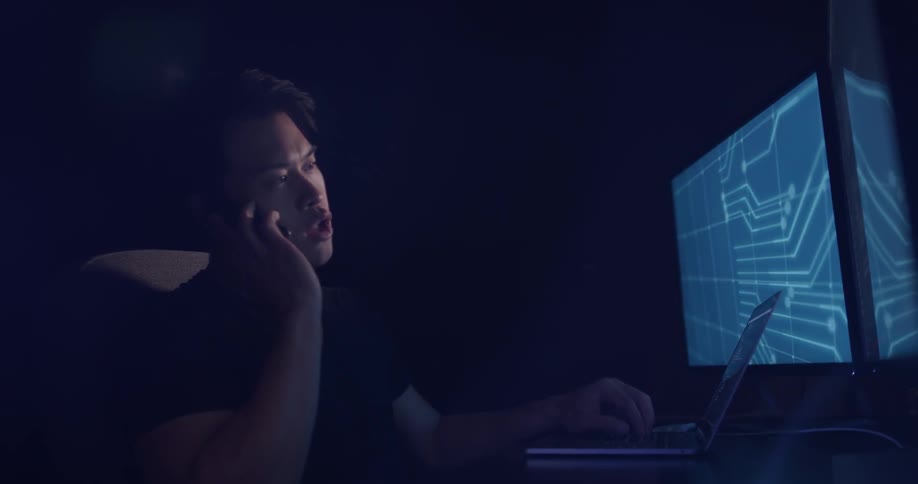 A man monitors computers and answers the phone in a dark room