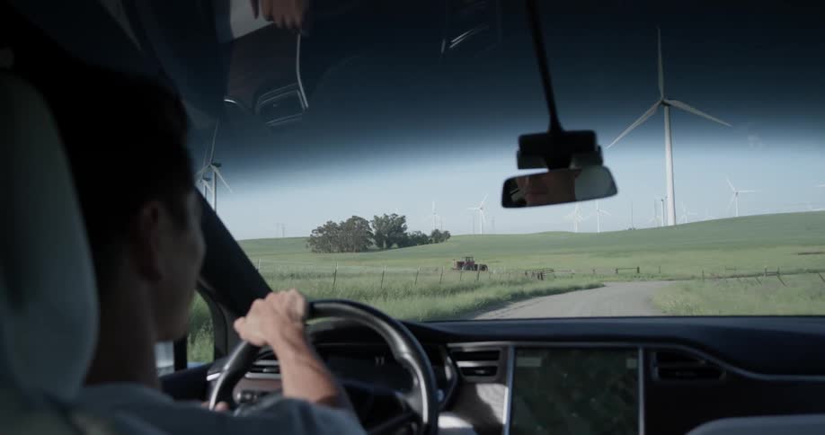Inside a car, a man drives on a country road towards a wind turbine