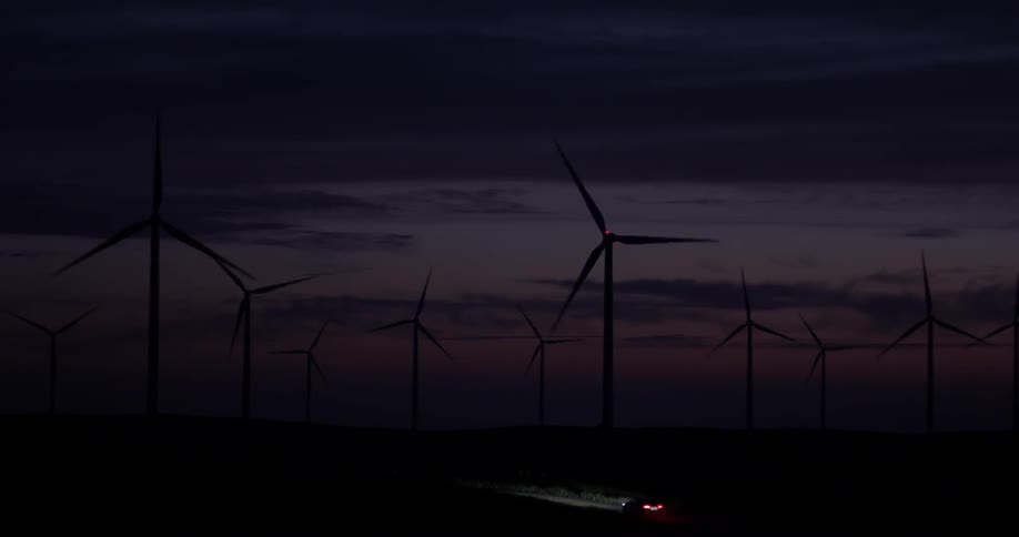 A windmill turns as a car drives below it in the dark