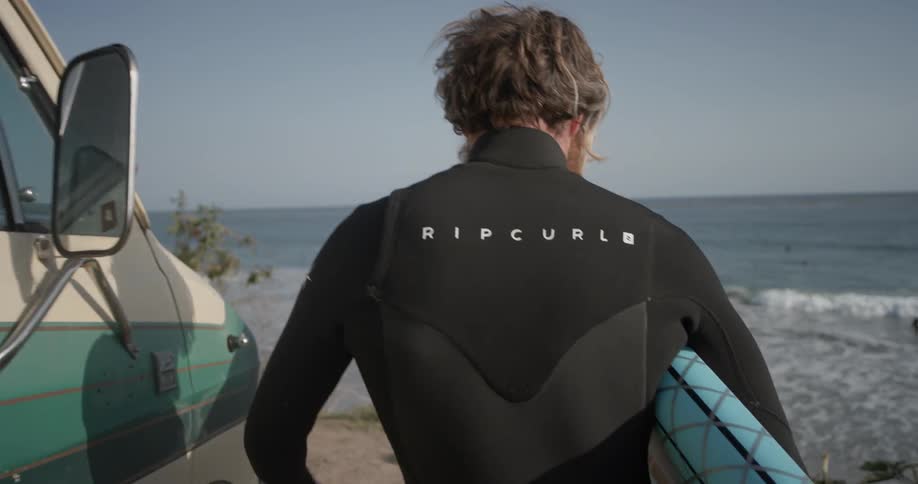 A bearded man walks past a van towards the ocean with a surfboard under his arm