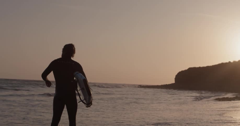 A surfer walks along the waterline on a beach with his board under his arm during a sunset