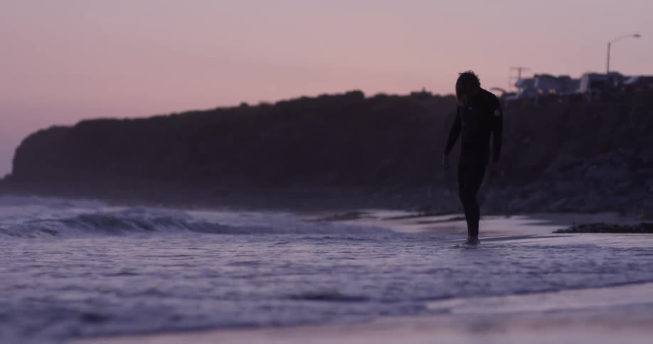 A man in a wet suit stands beside the ocean and begins to walk in the shallow water