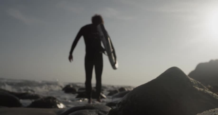 A man walking on rocks next to the ocean carrying a surfboard