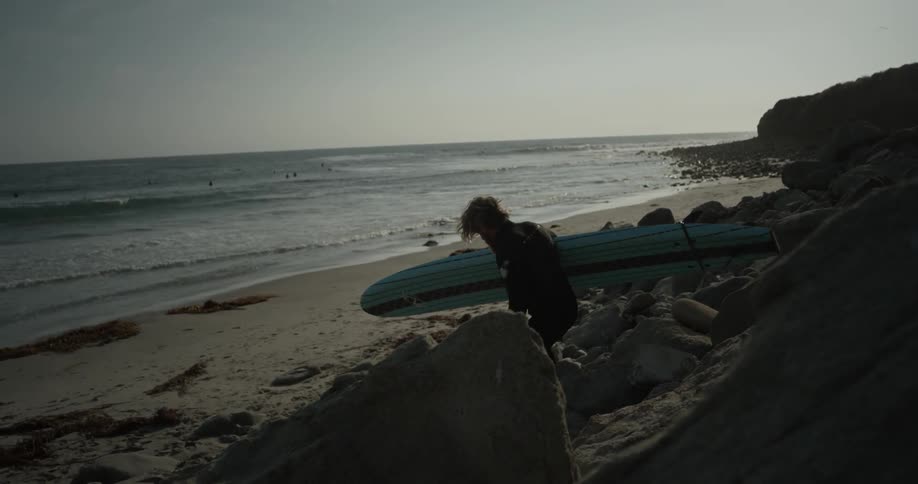 A surfer walks down rocks towards the beach with his board under his arm