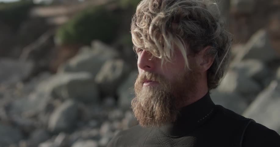 A man with scruffy hair and a beard wears a wetsuit