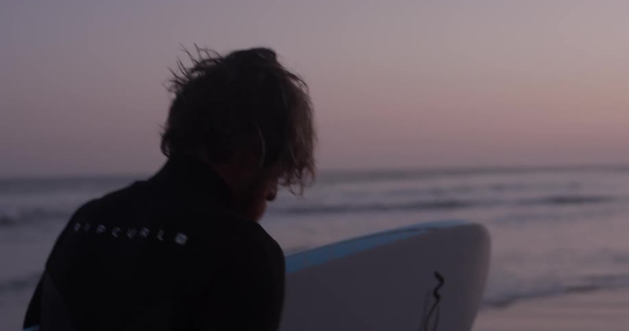A man in a wetsuit carrying a surfboard along the beach at sunset