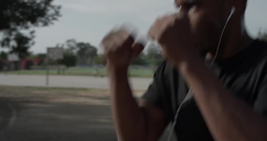 A black man is seen practicing his moves of boxing and wearing earbuds out at the park on a bright sunny day while he sweats from the exercise