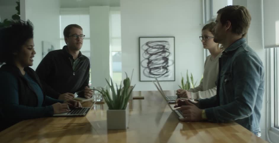 Four people talking at an indoor table with three of their laptops open