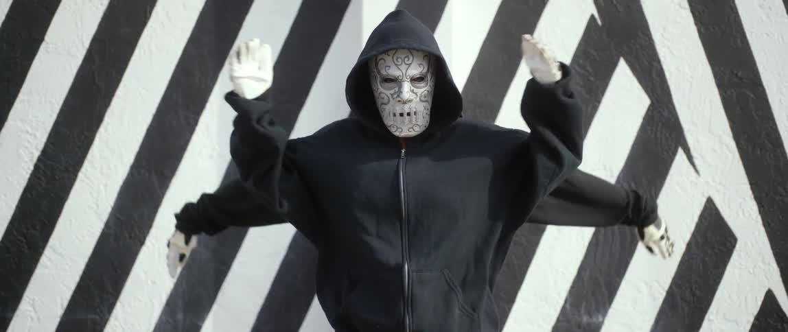 Two people, one is wearing a mask, dressed in black hoodies and skeleton gloves perform a synchronized dance in front of a black and white striped wall