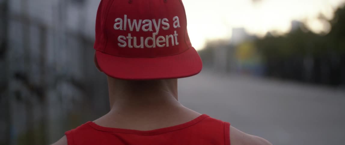 A man in a tank top and a backwards baseball cap walks down a street