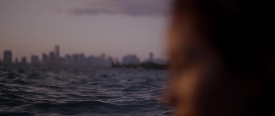 A woman is outside by a river and city buildings are in the distance