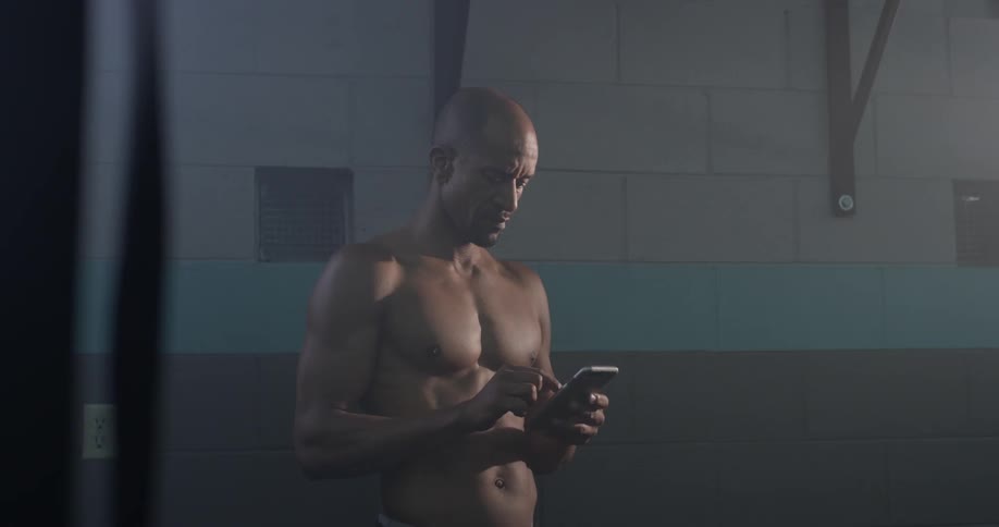 The top half of a shirtless man standing in a gym reading on his smartphone